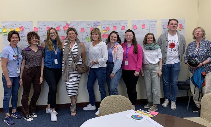 A group photo of the CCRT team standing in front of a series of posters with text and post-its. The posters represent the process mapping. Everyone is smiling.